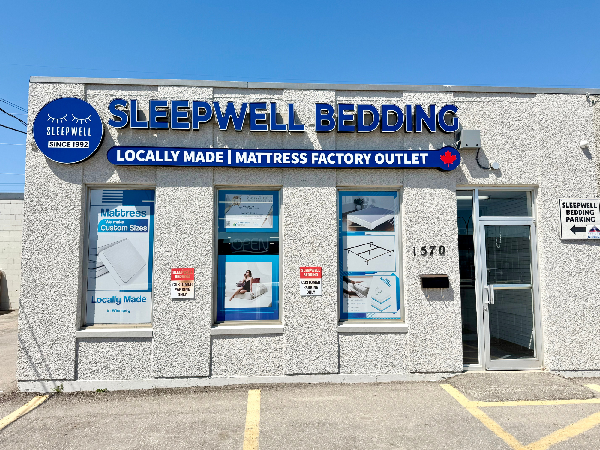 Front exterior of a Sleepwell Bedding store on a light gray stucco building under a blue sky. Large blue and white signage reads 'SLEEPWELL BEDDING,' with windows displaying mattress and bedding products and a parking sign on the right.